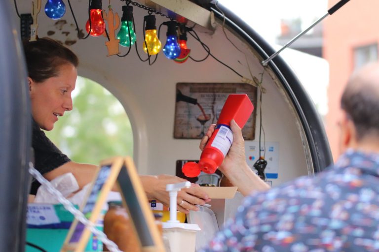 Stamtafel foodtruck Ilse Persoon serveert condimenten vanuit een kleine kraam met kleurrijke verlichting.
